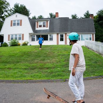 Young boy wearing a teal helmet with a foot on his skateboard that is standing on the street outside of a white Ädelbrook extension home