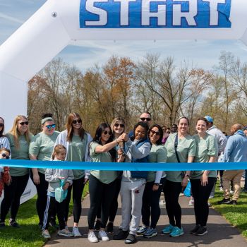 Large group of people  in front of an inflatable arch that says "Start" on it while one person cuts a blue ribbon with large scissors