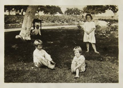 A vintage photo of 4 children playing outside on the grass, below a tree.