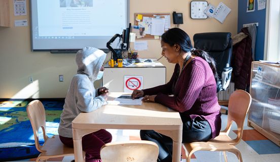 Woman sitting down and working with a young boy on a worksheet in a classroom