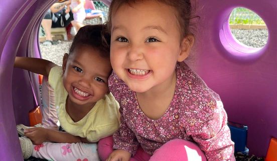 Two toddlers smiling while sitting in a playground tunnel