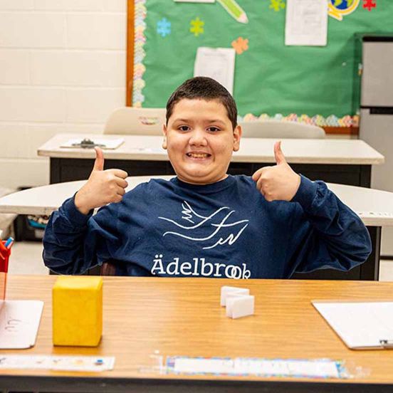 Young boy smiling with both his hands making a thumbs up, while sitting at a desk in a classroom 
