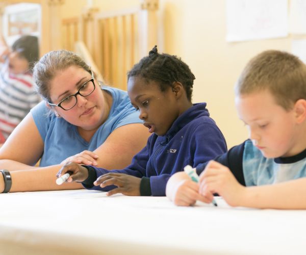 A woman helping a young boy who is writing with a marker with another boy writing in the foreground