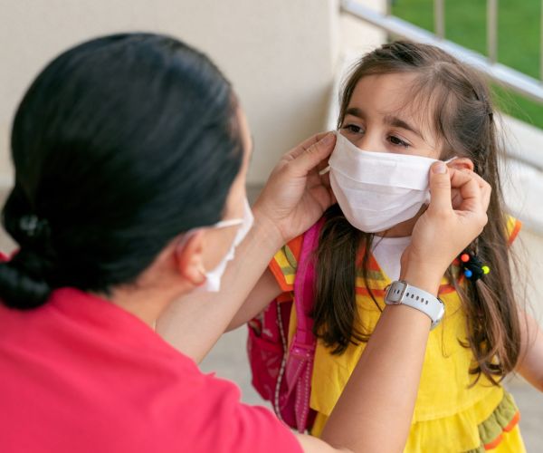 A child wearing a yellow shirt receives help putting on a white mask. 