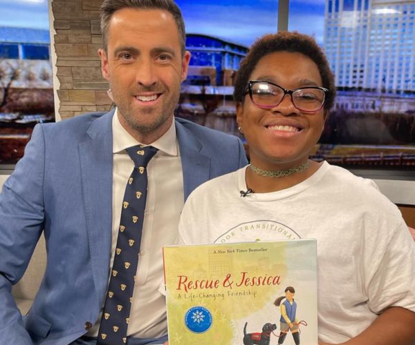 A news anchor smiling next to a girl holding a picture book called "Rescue & Jessica" while sitting on a couch
