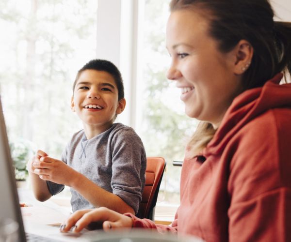 A smiling woman and child use a laptop together, with a bright window in the background