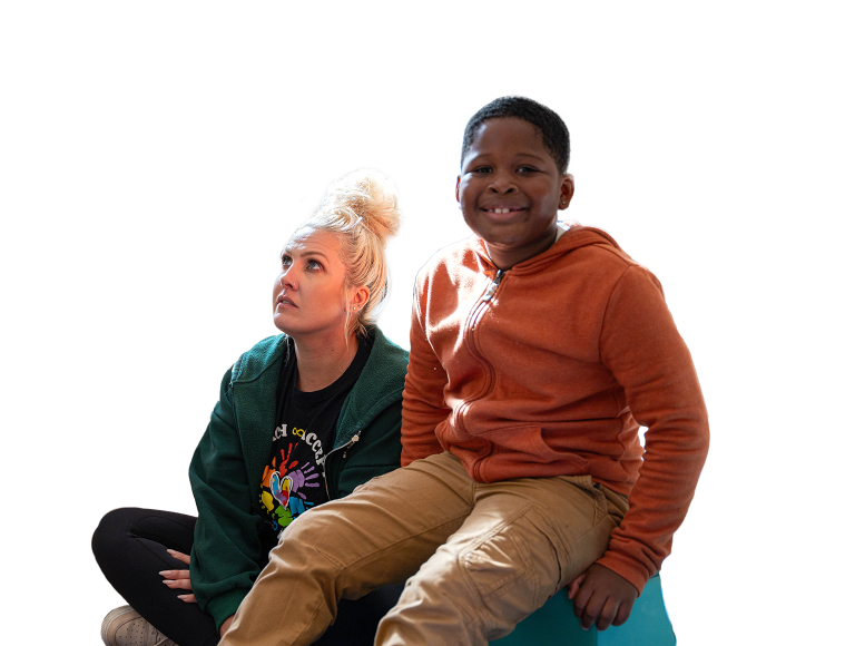 Young boy in an orange zip-up hoodie smiling for the camera, while an adult woman sitting next to him looks up into the distance