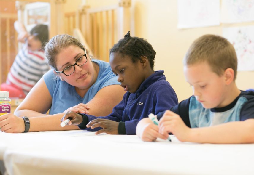 A woman helping a young boy who is writing with a marker with another boy writing in the foreground