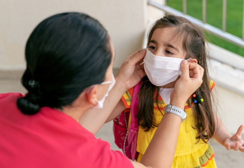 A child wearing a yellow shirt receives help putting on a white mask. 