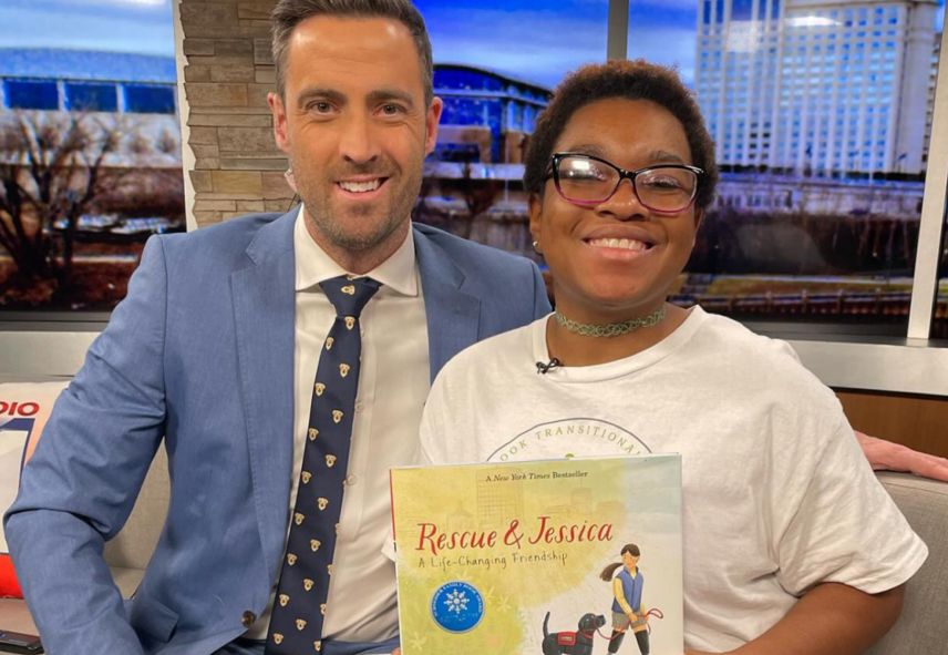A news anchor smiling next to a girl holding a picture book called "Rescue & Jessica" while sitting on a couch
