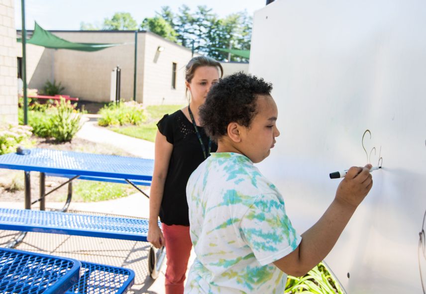 Young boy writing on a white board with an adult supervising him