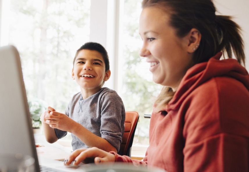 A smiling woman and child use a laptop together, with a bright window in the background