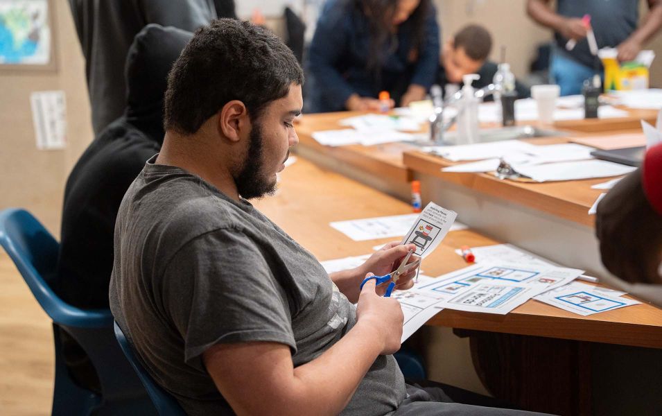 Young man sitting at a table and cutting out pieces of paper with scissors