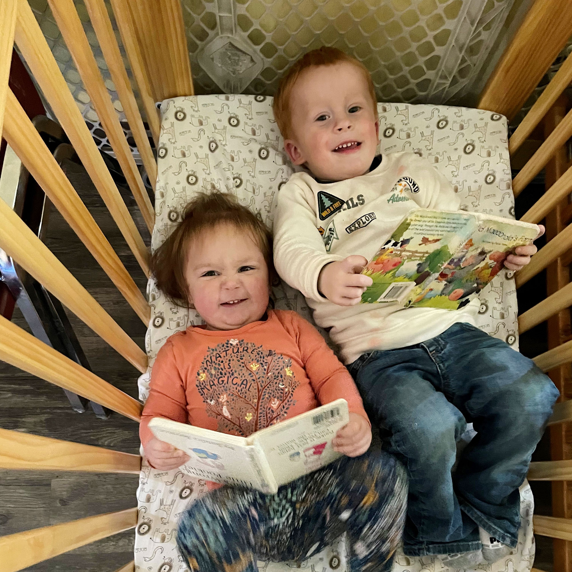 Two toddlers read books and giggle in a crib together.