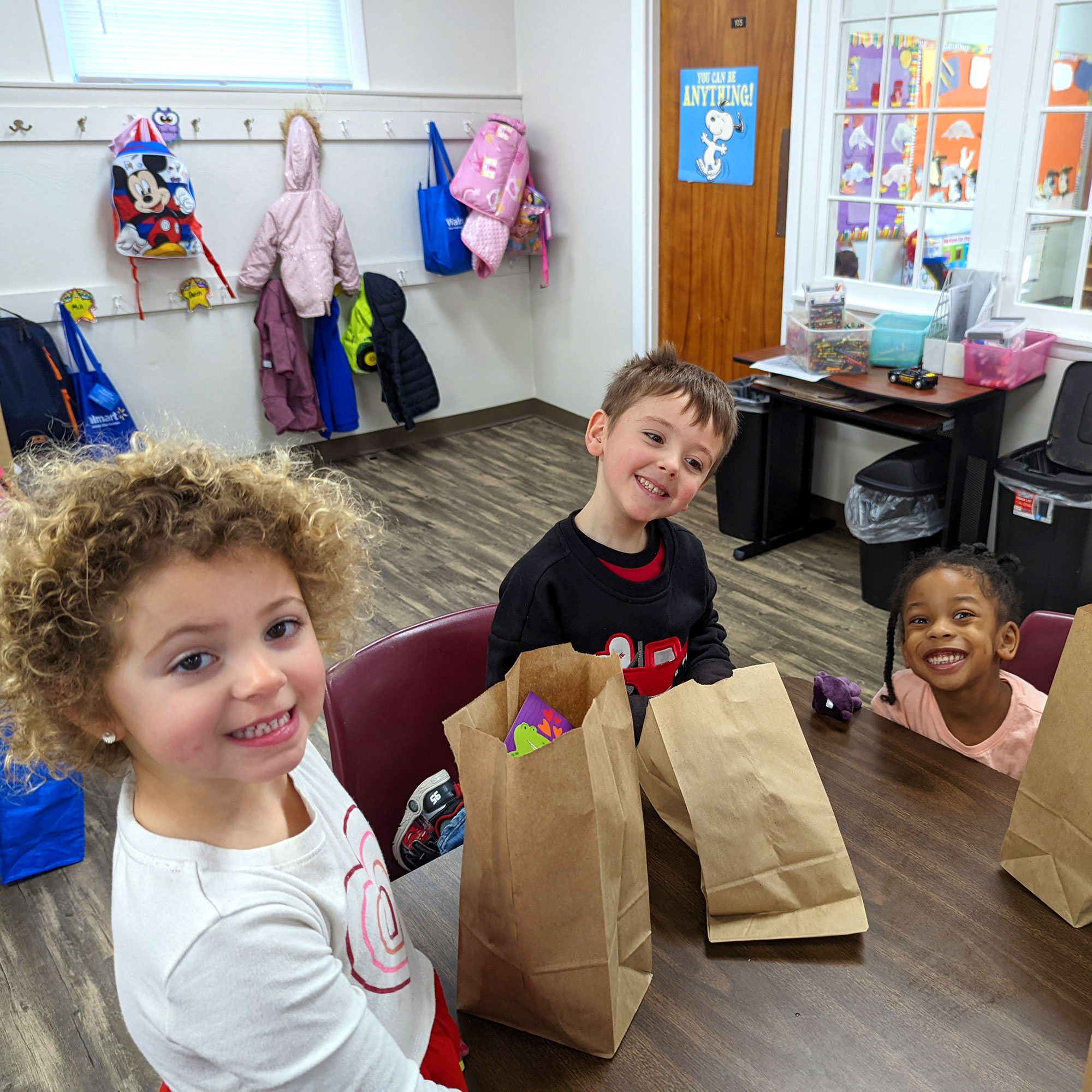 Three young children inspect their lunches in brown bags at school.